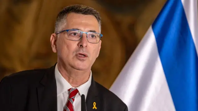 Gideon Saar speaking with the Israeli flag behind him.