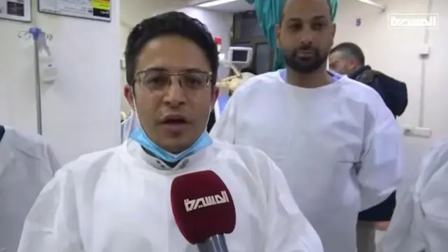 A doctor, wearing a white coat and with a blue fask mask around his chin, speaks to the camera. There's a hospital ward behind him.