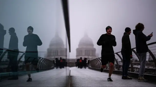 A jogger crosses The Millennium Bridge with the dome of St Paul's Cathedral disappearing into the fog behind. Other people stand by the rails of the bridge taking a photo