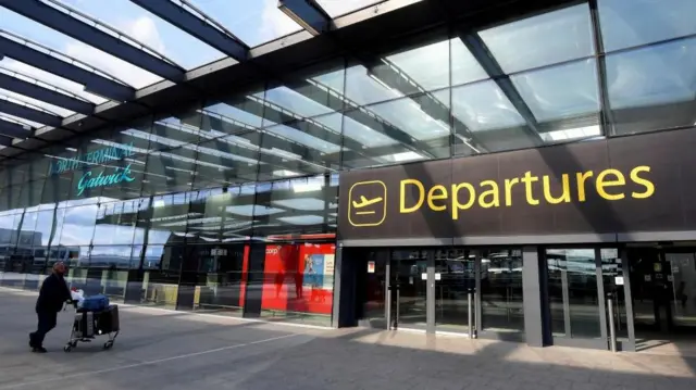 A man using a luggage trolley walks towards the airport entrance with a sign 'Departures'