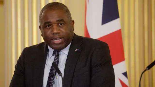 David Lammy in a dark gret suit and patterned tie sat in front of a white backdrop with a union jack flag, with a microphone in front of him