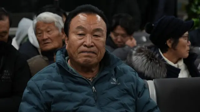 A man dressed in a blue puffer jacket identified as Maeng Gi-su sits with other bereaved families in the airport. He is frowning and looks very upset. Other people can be seen in the background with sombre expressions