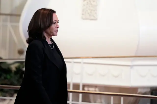 Kamala Harris in black suit walks in front of gold railing and white wall of church