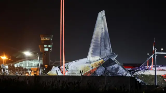 The wreckage of an aircraft that went off the runway and crashed, lies at Muan International Airport. Orange straps extend from what appears to be a door in the fuselage as workers attempt to lift the plane. The airport can be seen in the background and it is night time.