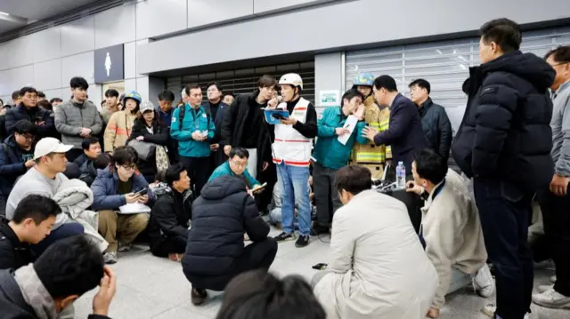 People gather around, some crouch on the floor, as an official speaks into a microphone