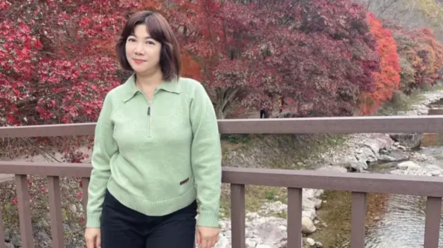 A Thai lady stands on a bridge in a green sweater and dark pants, with medium length hair, with red and pink trees and a stream in the background