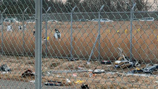 An image taken behind a metal gate. Forensics workers can be seen searching the ground. Debris lies on the foreground