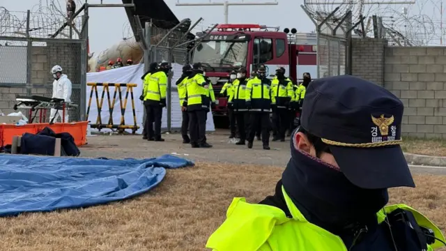 An emergency service worker stands in the foreground with his head bowed down wearing a cap. A number of other personnel in high-vis are in the background