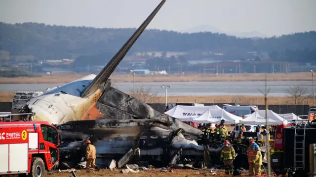 Firefighters stand next to a broken piece of plane that is lying charred on the ground. Tents are nearby