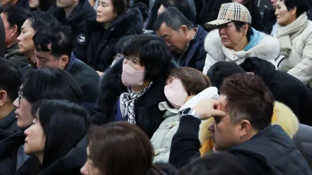 A crowd of people look ahead. Some appear visibly distressed. Two women in the centre of the image wear pink masks. One rests her head on the other's shoulder