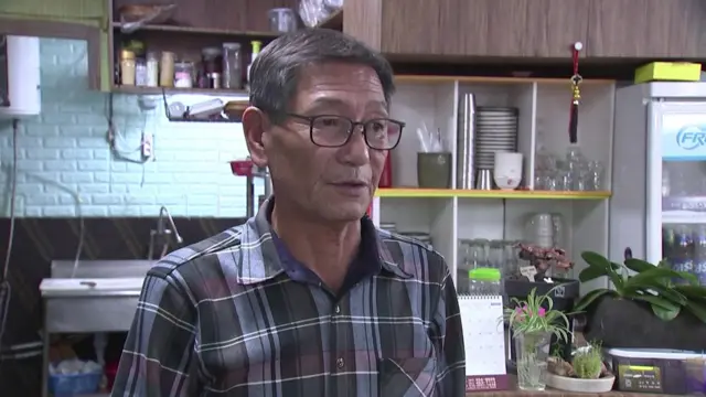 Im Young-Hak wears a checked shirt. He looks to the right of the camera. In the background are shelves with plates, glasses and condiments