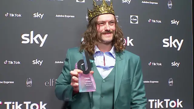 Iain Ward wears a crown and green suit as he holds his award