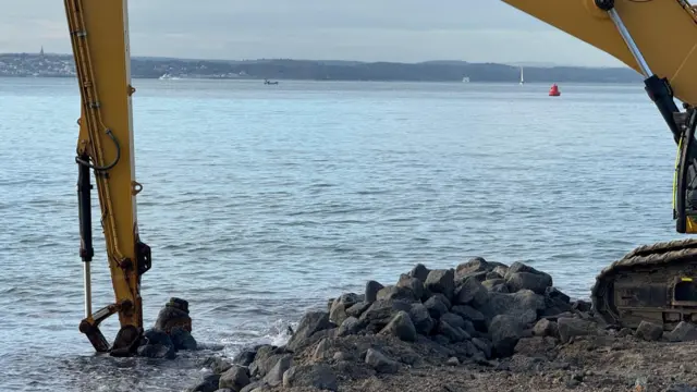 A digger with its arm in the sea next to a small stack of rocks. A metal cylinder can be seen poking out of the water just next to where the digger was digging.