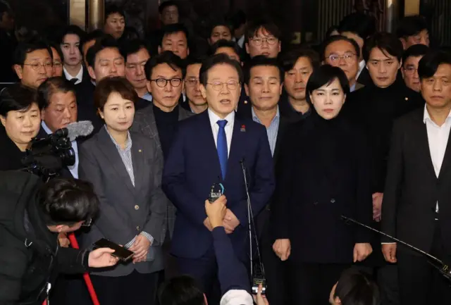 South Korea's main opposition leader Lee Jae-Myung speaks with a microphone in front of him as he speaks to the media. There's a crowd of people standing behind him.