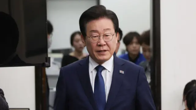 Democratic Party leader Lee Jae-myung walks out of room. He's wearing a dark blue jacket with matching tie and a South Korea flag pin on his left lapel