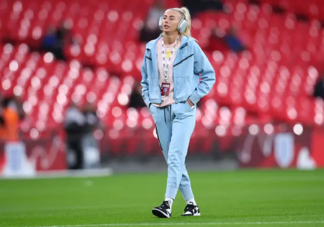 Chloe Kelly in a tracksuit walking on the pitch pre match with headphones on