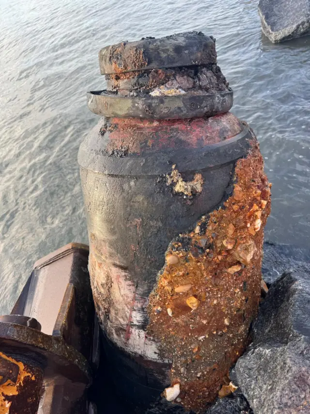 A large metal cylinder with rock stuck to one side of it. The sea and the arm of a digger can be seen behind it.