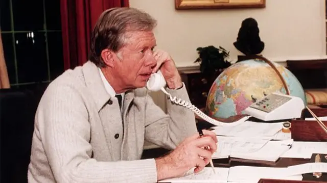 Jimmy Carter, pictured in 1980, dons a sweater in the White House. He's sitting at a desk and answering a call.