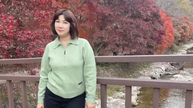 A woman in a green jumper on a bridge with trees and a river behind her