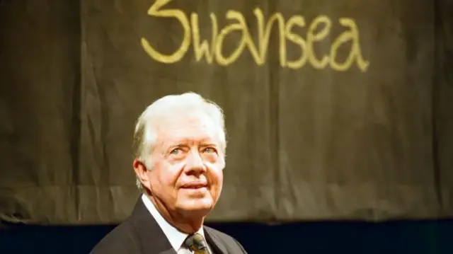 A photo of Jimmy Carter from the shoulders and up, with a black sign saying Swansea in the background.