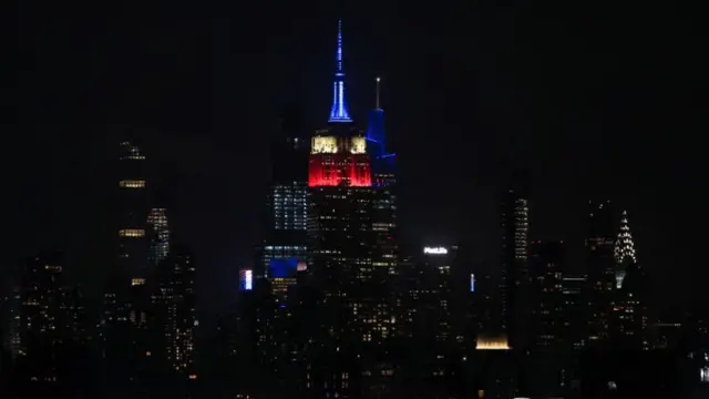 A shot of the New York skyline at night. In the centre, the top of the Empire State building is illuminated in blue, white and red against the dark sky