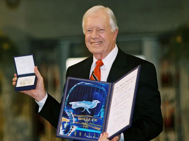 Carter, wearing a suit, smiles as he holds up a small box containing the medal and a certificate