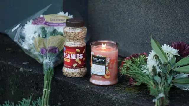 Between two bouquets, a jar of peanuts and a peach-coloured candle sit on a small, black stone step at the base of a sign