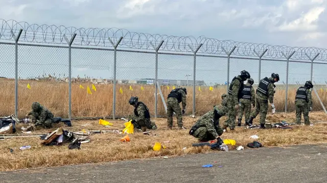 Group of soldiers searching and scanning grass covered in plane debris next to fence with barbed wire