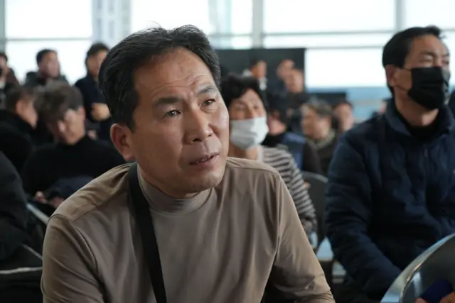 Man sat an airport, looking toward the camera in a beige shirt, with other people sat behind him blurred in the background