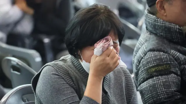 A woman in a grey jumper sits on a chair holding her hand with a handkerchief in it covering her face, visibly upset