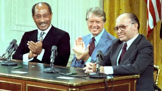 The three men applaud as they sit side by side at a desk in front of a gold curtain and US flag