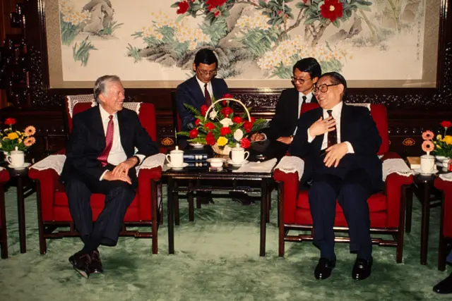 Jimmy Carter meets Jiang Zemin, who was the Chinese Communist Party Secretary at the time, in Beijing in 1991. They're both sitting in red chairs, with a table with tea and a bouquet of flowers on it.