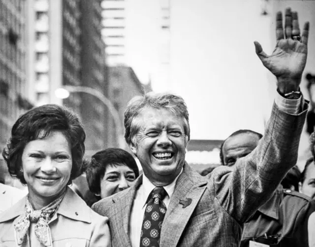 A black and white photo of Jimmy Carter waving to a crowd while standing next to his wife Rosalynn