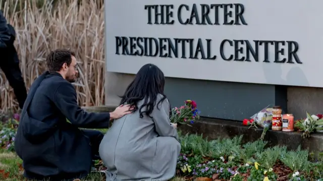 A woman crouches by a large sign that read The Carter Presidential Center, holding a small colourful bouquet. A man beside her rests a hand on her back