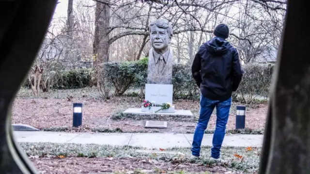 A man stands off a path, looking at the large stone bust at the base of which are bouquets of flowers