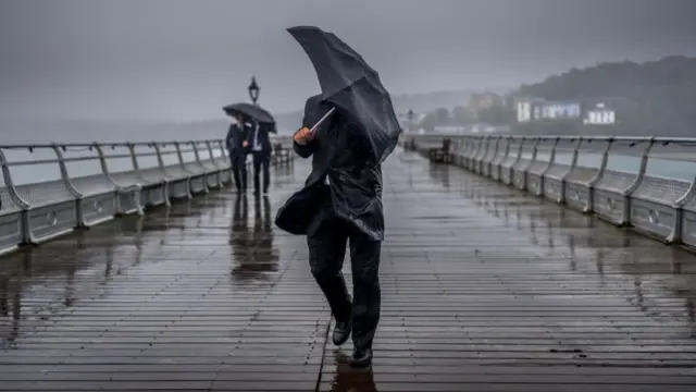 Man in black rain jacket battles against the wind as he covers himself with a black umbrella while walking down Garth Pier in Bangor. Two other men in suits walking under an umbrella are behind him, a small selection of buildings visible in the far right of the frame