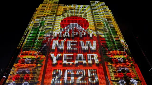 A skyscraper is lit up with a graphic of 'Happy New Year' on top of a snowy mountain and a rising sun