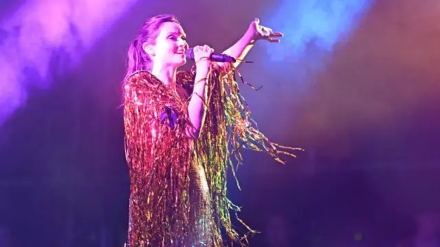 Sophie Ellis Bextor in silver sparkly dress with orange fringe fabric cascading down off her shoulders, with microphone in her hand and one arm lifted with finger pointing up