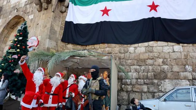 Four men dressed as Santa take a selfie with a fighter of the ruling Syrian body, who is holding a gun and wearing a black balaclava