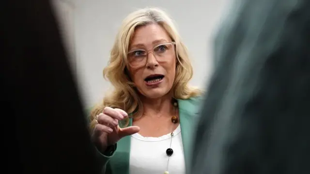 Gilliam Martin mid-speech with her right hand raised as she looks to her left. She's wearing a white t-shirt and pale turquoise blazer, a white, black and brown stone and silver string necklace around her neck. In the foreground are two blurry arms covered in shirts