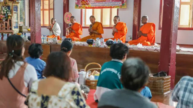 A line of Buddhist monks are seated in orange robes in front of a group of villagers as they pray