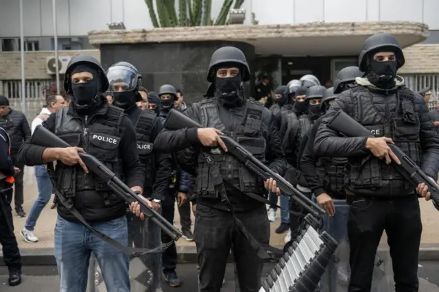 A group of policemen lined up wearing helmets, face coverings and holding large guns