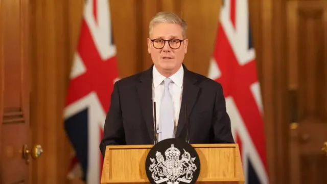 Keir Starmer wearing black glasses and a suit, standing at a lectern speaking into a microphone. Two British flags are behind him