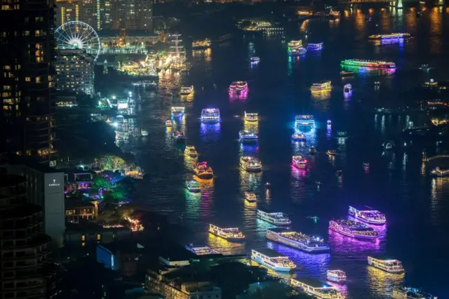 Lots of large boats on a river. They are all lit up in different bright colours. Skyscrapers border the river, and a big wheel is seen in the distance