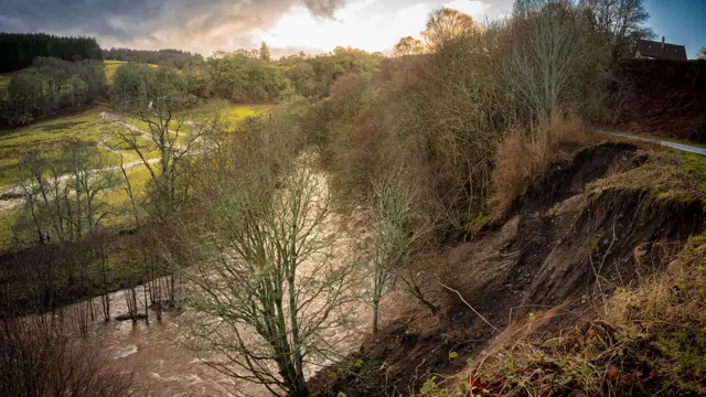 A dramatic landscape of a flooded field, a raging brown river, and a muddy landslip