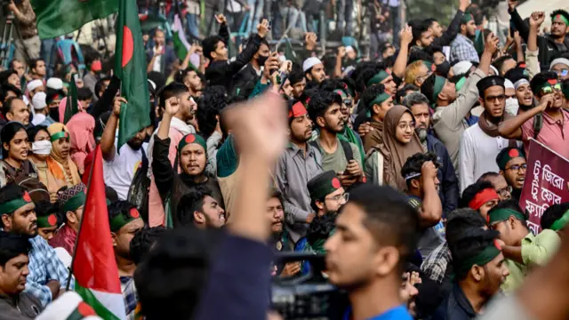 A large gathering of people, some with their fists in the air, some holding flags and others wearing headbands with the Bangladeshi flag, protest in Dhaka.