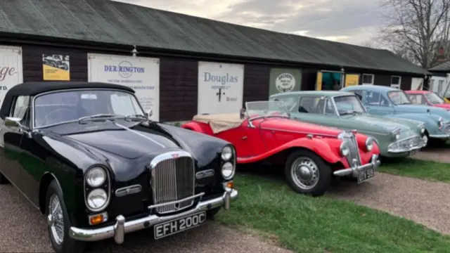 Line-up of vintage cars parked outdoors in front of a brown shed. From left to right: a black car, a red convertible, a pastel green car, a light blue car and a red car