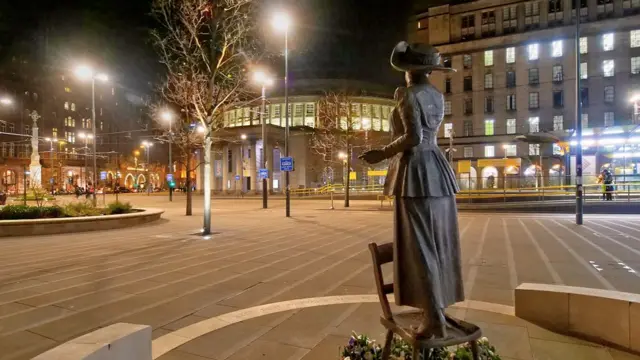 A deserted city square with the statue of a woman who is on top of a chair facing away from the camera. There are a few bunches of flowers at the feet