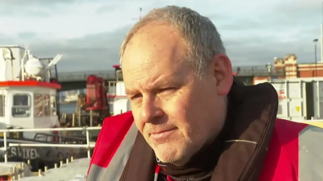 Darryl Fleming close up cuts off below the shoulders. He's wearing a red hi-vis vest as he stands next to the river on the embankment. A small boat is visible behind him to the right, a red and white brick bridge is also partially visible in the background