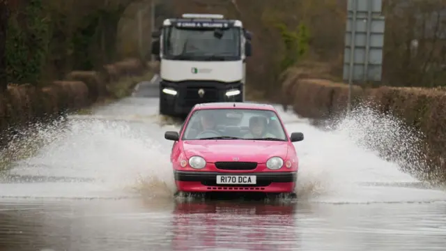 A small red car driving through a flood sending water shooting up on both sides. A lorry is behind it. A line of brown shrubbery lines up the road on both sides